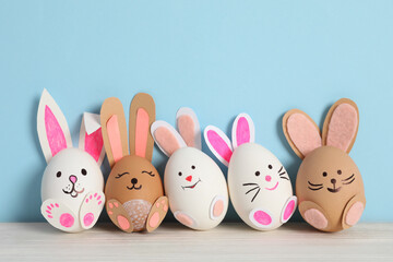 Eggs as cute bunnies on white wooden table against light blue background. Easter celebration