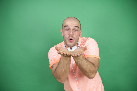 Young bald man using headphones over green background Smiling with hands palms together receiving or giving gesture. Hold and protection