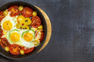 Fried eggs in a frying pan with cherry tomatoes and bell pepper for breakfast on a black background.