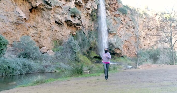 Dron volando y mujer corriendo con mascarilla covid-19 junto a paraje con catarata de agua