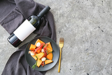 Plate with delicious melon, mozzarella, prosciutto and bottle of wine on gray background