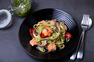 Noodles with seafood, sun-dried tomatoes, capers and red onions. Homemade pasta with shrimp, salmon (trout) and pesto sauce. Black background, black plate. Close-up.