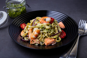 Noodles with seafood, sun-dried tomatoes, capers and red onions. Homemade pasta with shrimp, salmon (trout) and pesto sauce. Black background, black plate. Close-up.