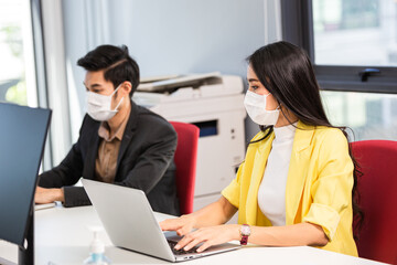 Asian business woman and colleague wearing surgical mask working with laptop in office. new normal lifestyle at workplace. company reopen during coronavirus spreading. people keep social distancing. 