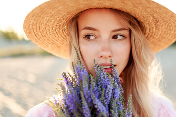 Close up lifestyle portrait of romantic blonde woman with flowers in hand walking on sunny beach . Warm sunset colors.