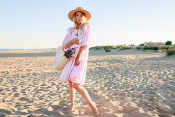  Full height  image of   blond   girl in cute pink  dress dancing and having fu on  the beach. Holding straw bag  and  flowers.