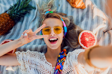 Top view on pretty  blond  laughing  woman lying on beach towel, having fun, spending great time . Tropical fruits, pineapple. Boho accessories.  Shows signs by hands.