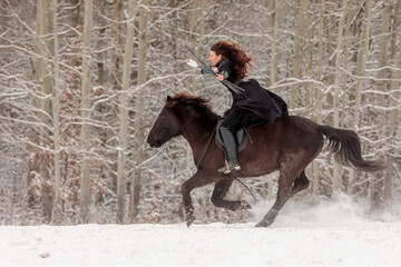 young woman riding in historical costume as a warrior and rides through a winter landscape with a bow in her hand