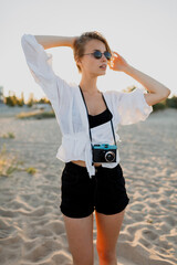 Elegant blond woman  with retro camera making grimaces and posing on the beach near ocean. Summer holidays. Beautiful sunlight.