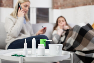 Close up of medicines, tablets, spray and drops for domestic treatment of flue or virus infection. Blurred view of woman calling to the doctor, checking her sick daughter's temperature laying in bed