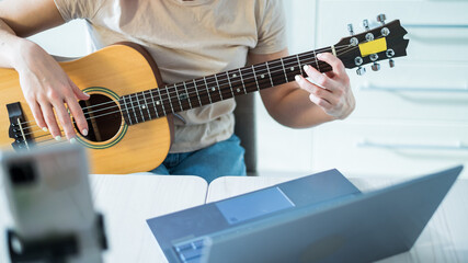 A woman sits in the kitchen during a remote acoustic guitar lesson. A girl learns to play the guitar and watches educational videos on a laptop