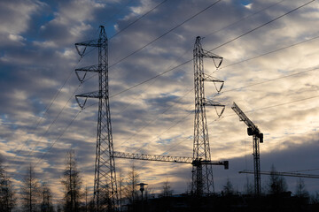 A tall construction crane against the setting sun. High voltage power pole. Machine for high-altitude lifting of building materials. The tops of conifers.