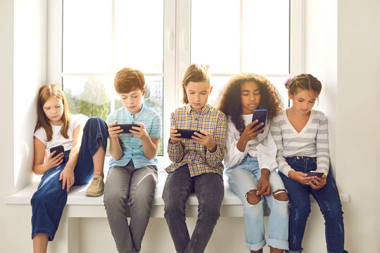 Multiethnic group of 8-10 year old children sitting on windowsill, playing online phone games and ignoring real life. Concept of gadget addiction and excessive use of social media and mobile devices