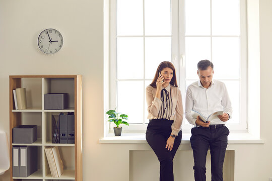 Two Young Business People Standing By The Window In Office Workspace During Break At Work, Man Is Looking Through Papers, Woman Is Talking On Mobile Phone