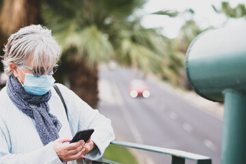 Coronavirus. Senior woman with cellphone in the hand reading a message. Elderly retiree standing on an elevated city street wearing surgical mask due to coronavirus.