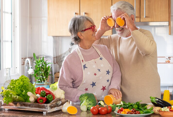 Beautiful elderly couple in the kitchen joke laughing with two slices of pumpkin. Joyful healthy elderly lifestyle. On the table a mix the of raw seasonal vegetables. Vegetarian lifestyle