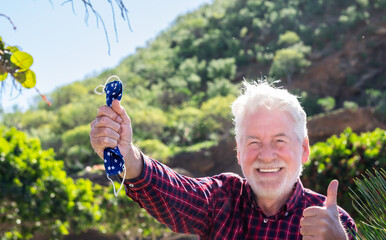 Blurred portrait of a senior man holding a protective coronavirus mask. Focus on hand and mask. He laughs looking at camera gesturing a positive sign. A joyful elderly person with white hair and beard