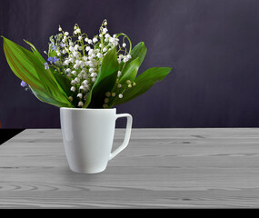 Spring still life with delicate lilies of the valley and forget-me-not flowers in white cup on wooden table. Bouquet of lily of the valley flowers on dark background in vintage rustic style, space for
