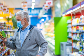 Adult senior man with surgical mask due to coronavirus while shopping at the supermarket. White haired retiree with shopping cart, choosing a canned product. Consumerism concept