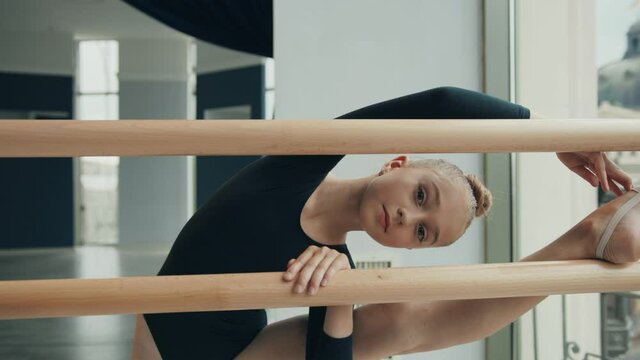 Cute little girl in black sports swimsuit stands in studio with one leg thrown on ballet machine and leaning over looking at camera smiling.
