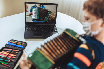 Focused boy playing accordion guitar and watching online course on laptop while practicing at home. Online training, online classes.