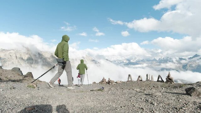 Group of hikers dressed in warm ski suits walking towards edge cliff with white fluffy mountains. Clip. Female name made of stones and travelers men and women walking on a stony flat surface with ski