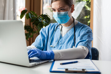 Young caucasian female doctor working on laptop computer in office, wearing protective glove and face mask