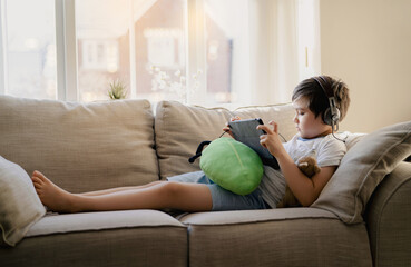 Authentic portrait Kid sitting on sofa watching cartoons on tablet,Yong boy playing game on touch pad, Child lying on couch wearing headphoes listening to music or relaxing on his own in living room