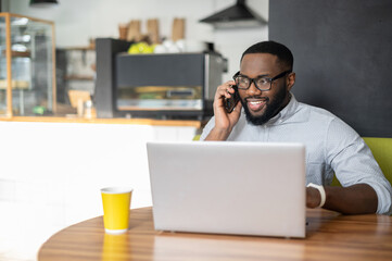 Smiling African-American guy is using a laptop for remote work sitting in the cozy cafe. A multiracial freelancer is talking on the smartphone sitting at the desk with a computer in coffee shop