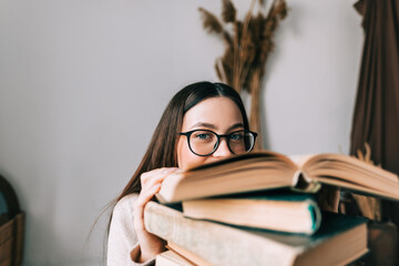 Portrait of young caucasian woman college student in eyeglasses hiding behind a stack of books and...