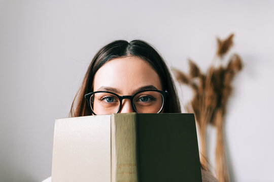 Portrait of young caucasian woman college student in eyeglasses hiding behind a book and looking at camera.