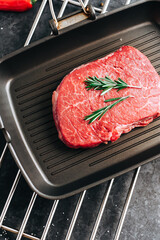 Raw beef steak on black grill pan with rosemary, top view.