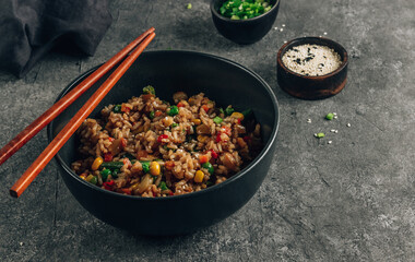 Fried Rice with vegetables, green onion and sesame in black bowl on stone background.