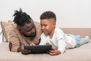 Fototapeta premium Dark skinned African male and his little son lie on the sofa at home and use tablet