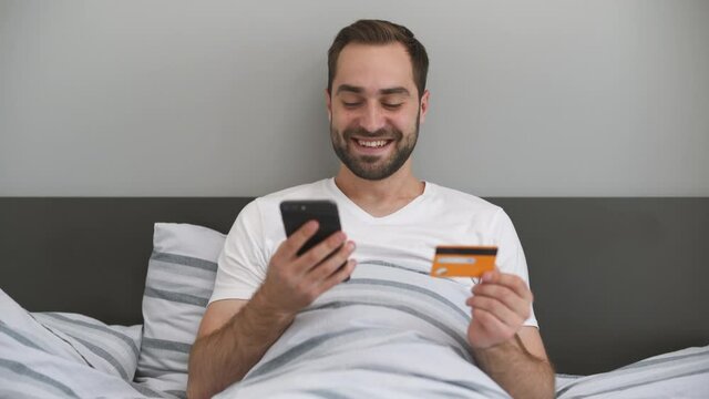 Cheery young man in white t-shirt lying in bed rest relax spend time in bedroom lounge home in own room house hold in hands credit bank card using mobile cell phone order delivery. Real estate concept