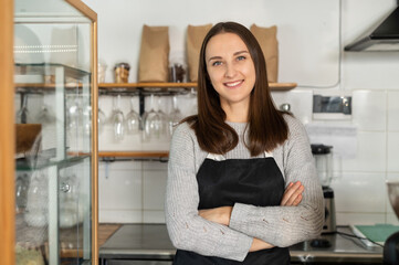 Confident female business owner standing at the counter of a restaurant with arms crossed, bakery waitress in apron looks at the camera and smiles