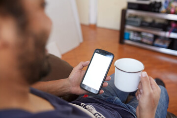 point of view hispanic man sitting on his living room couch with a cup of coffee in his hand and browsing on his smartphone