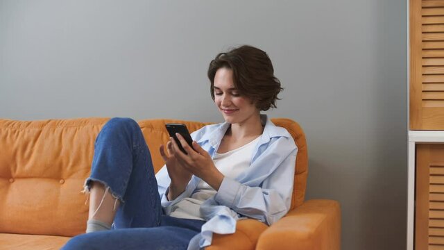 Cheery young woman in shirt sit on sofa couch rest relax spend time in living room lounge home in own house hold using mobile cell phone chatting serfing browsing typing sms dream Real estate concept