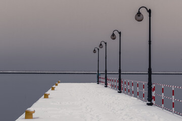 Photo of the pier covered with snow. In the foreground there are railings and lamps in the...