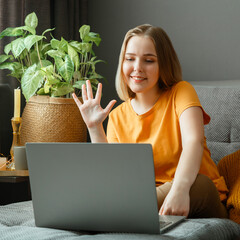 Happy young beautiful woman greets, talking by video call using laptop while sitting on sofa in living room. Remote work study, communication with family and friends in home interior