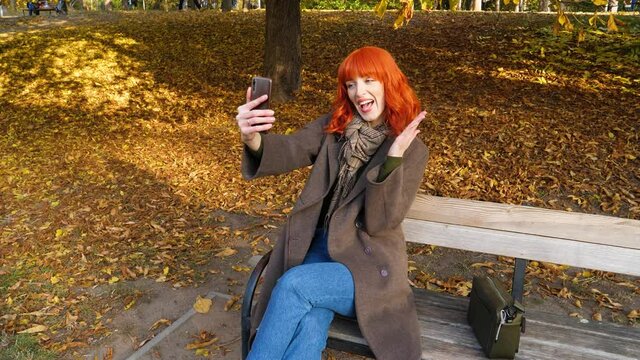 a girl with red hair sits on a bench against the background of yellow autumn foliage, and takes pictures of herself with different hand gestures and unusual facial expressions,