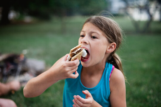 A tween bites into a messy s'more around a backyard campfire