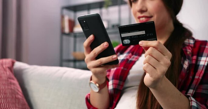 Let&rsquo;s buy this one. Young brunette woman sitting at the sofa at home, holding credit card and doing online shopping holding her cell phone. Impulse buying, online shopping, fast fashion, shopaholic