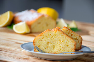 Piece of lemon cake on plate on rustic wooden board with full pie and lemons on background. Easy recipe of citrus dessert for everyday cooking. Homemade bakery by classic recipe.
