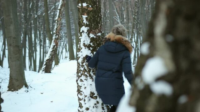 Portrait of a cute man peeking from behind tree. Woman hiding. Smiling attractive girl peeking out from behind trunk. Blind man's buff game, couple run after each other on snowy winter day