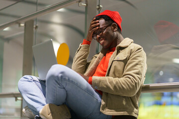 Surprised African hipster man wear red hat and glasses sitting on the floor in airport, talking in...