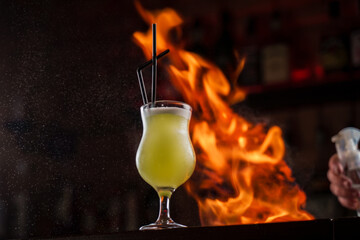 Bartender sprinkles on illuminated glass with bright green cold cocktail on bar counter and makes fire flame over it.