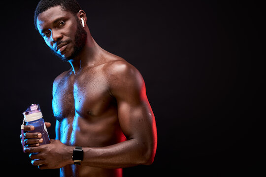 athlete black male with naked torso holding water bottle in hands, looking at camera, wearing wireless headphones