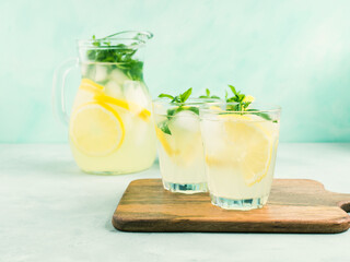 Fresh lemonade in jug and glasses with ice, mint leaves and lemon slices on bright gray and blue background. Closeup