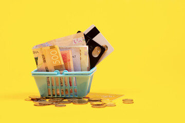 Shopping basket with coin, credit card and ukrainian bills hryvnias on yellow background. Space for text. Close up. Business concept.